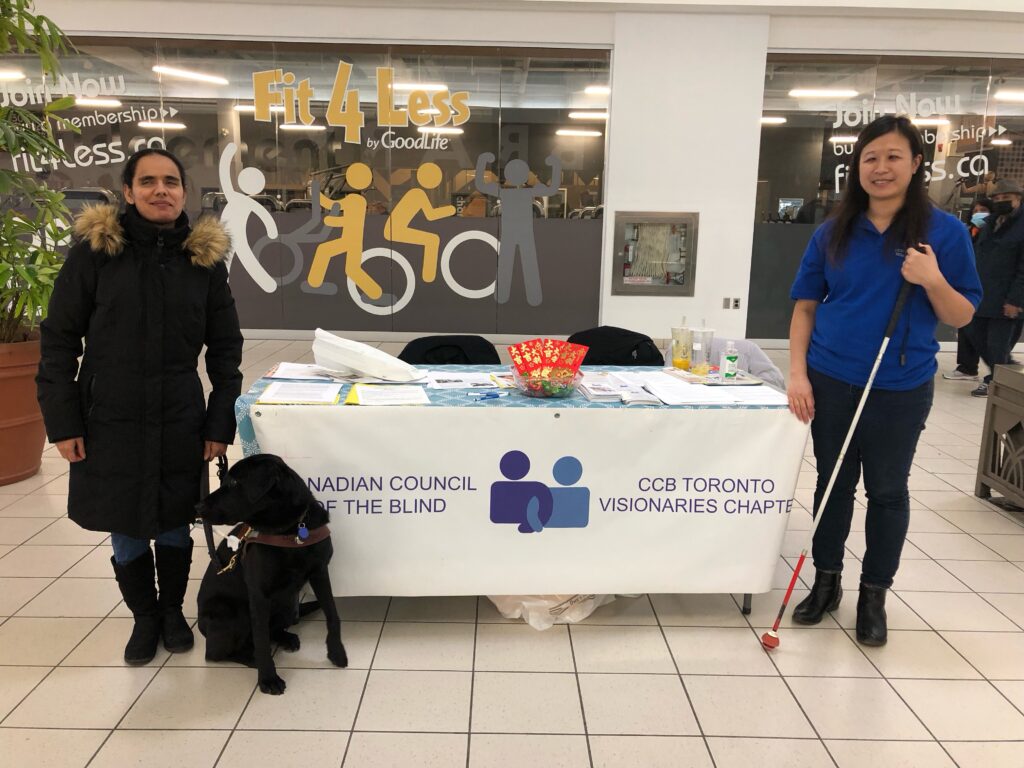 In the middle is  table with the CCB banner spread across with Wendy and
white cane to the right and Runa with Guide dog Steller to the left.